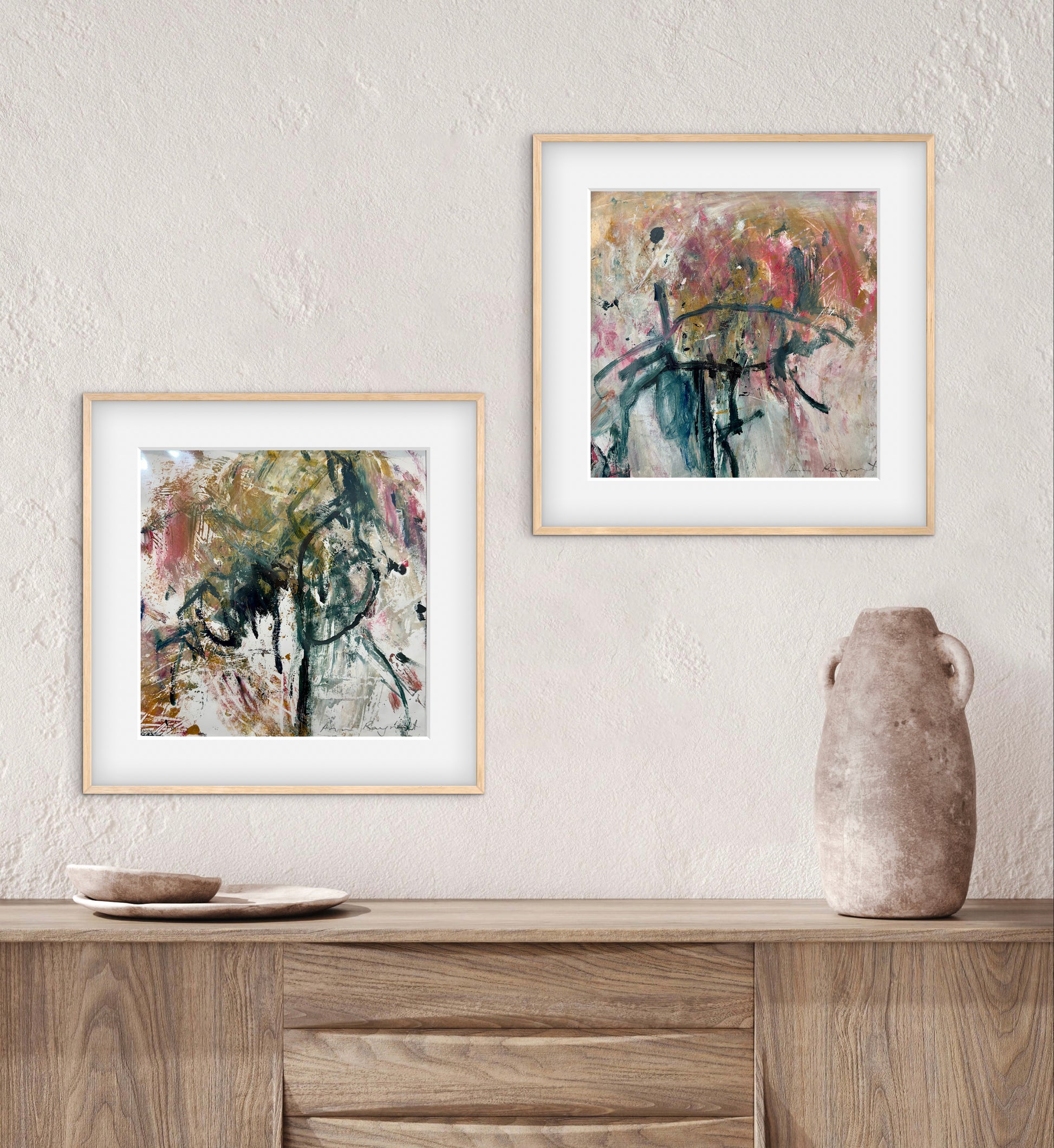Two abstract artworks in gold frames on a wall above a wooden surface with a vase.