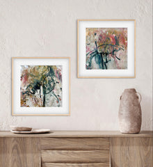 Two abstract artworks in gold frames on a wall above a wooden surface with a vase.